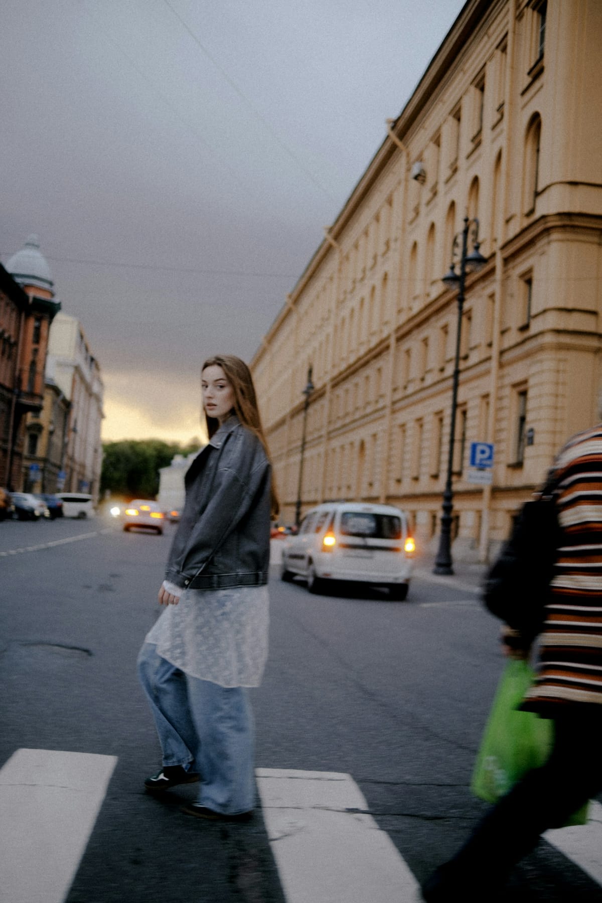 A woman crossing a city street at dusk, glancing back over her shoulder