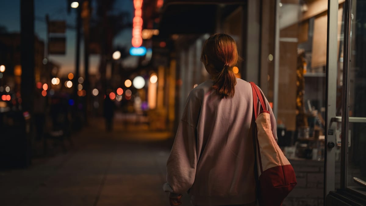 A woman with a tote bag walking home along a lit city street in the evening